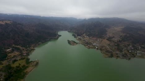 Vista-Aérea-De-Un-Lago-Sinuoso-Rodeado-De-Colinas-Y-Comunidades-Rurales-Bajo-Un-Cielo-Nublado-Con-Una-Pequeña-Isla-En-El-Lago