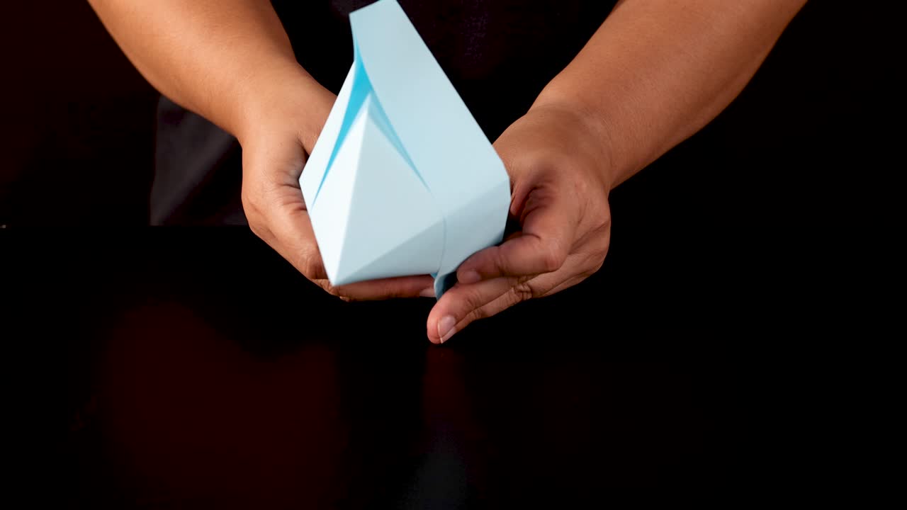 Hands carefully fold blue paper into an origami boat on black background, overhead lighting