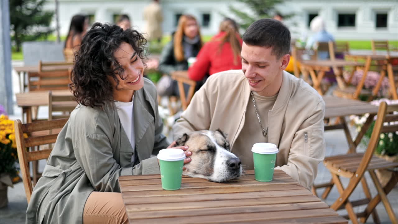 A happy couple outdoors near a cafe. Petting their dog, coffee on the table. Autumn atmosphere. Slow motion