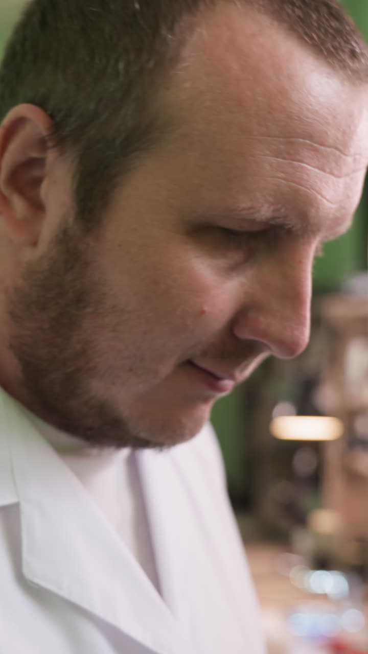 una vista en primer plano de un hombre con una bata blanca de laboratorio caminando dentro de su laboratorio, con un estante detrás de él con herramientas de trabajo en el estante y la pared pintada de verde