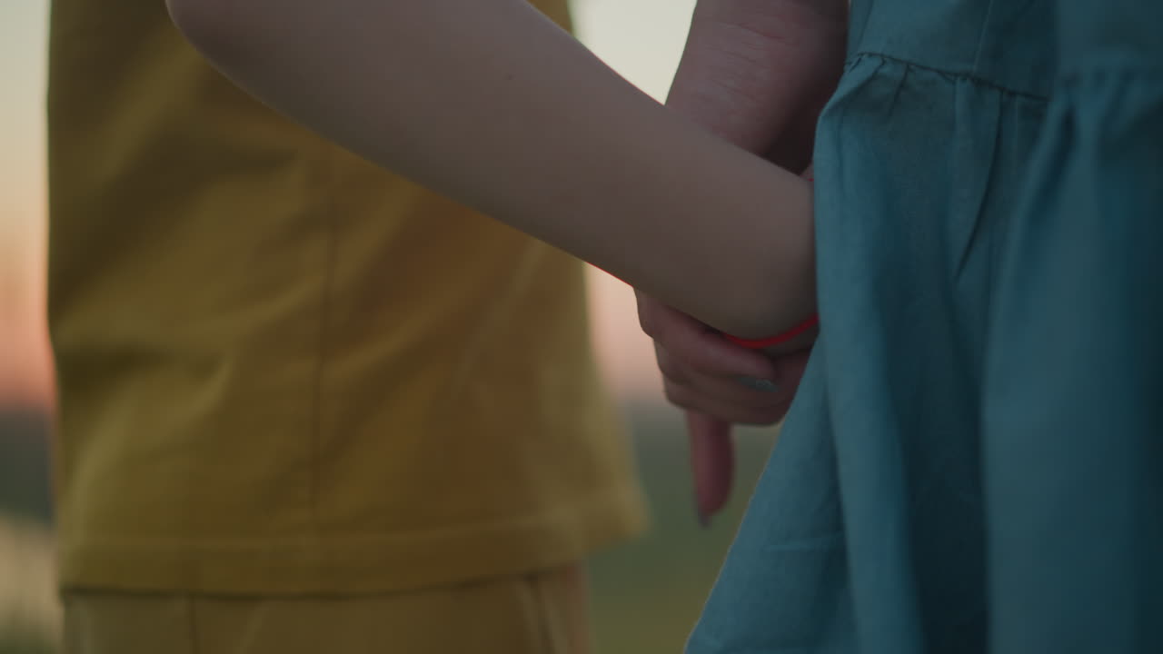 A touching close-up of a mother holding her son's hand, who is wearing yellow, as they stand together, facing away from the camera, watching the serene sunset over a tranquil lake