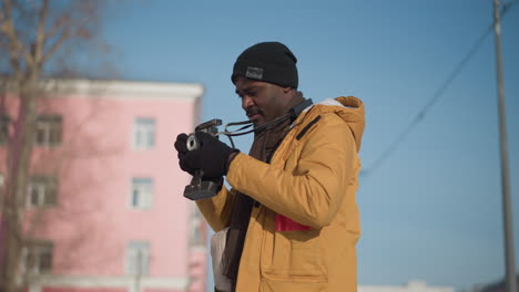 black photographer walking along snow covered city walkway with camera hanging on neck, holding camera out and looking around both sides amid blurred tall buildings under bright winter sky