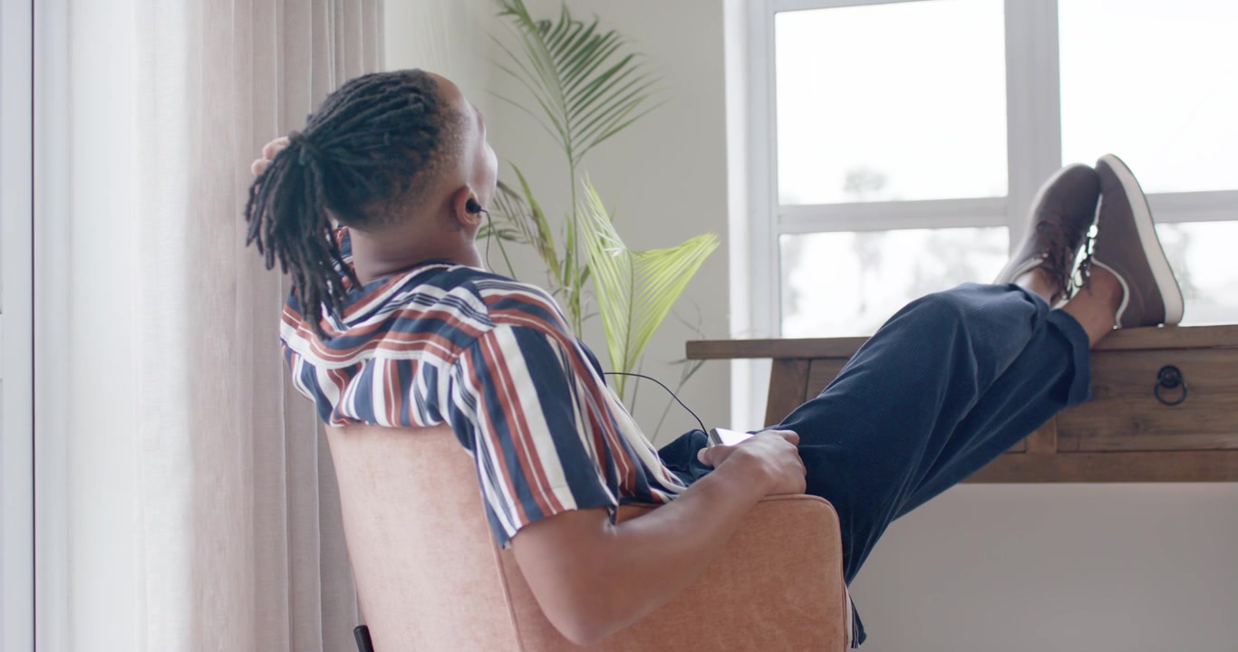 African American man reclining in armchair at home, shifting feet on desk for comfort near window