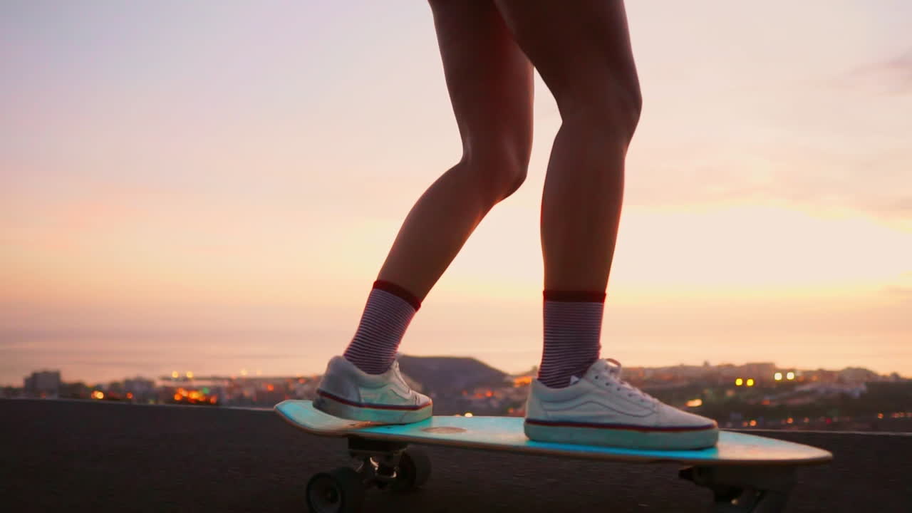 A mountain road serves as the stage for a stunning and stylish skateboarder, wearing shorts, as she rides her board during sunset, with the mountains' view emphasized in slow motion