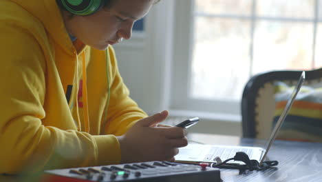 teenage boy on an iphone and headphones while sitting at a laptop with midi keyboard at table