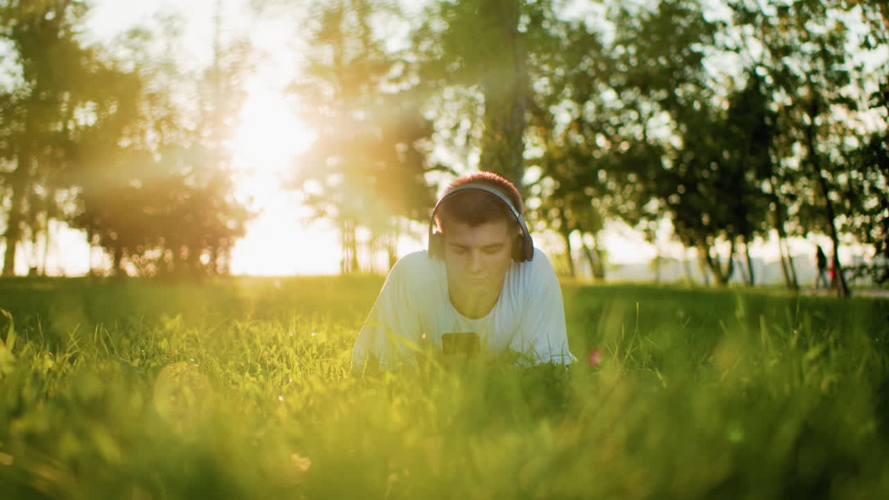 잔디 위에 누워있는 검은 헤드폰을 착용 한 젊은이가 빛으로 드폰에 초점을 맞추며 을 가로질러 따뜻한 빛을 만니다. 배경에는 울창한 초록색과 멀리 걸어가는 사람들이 있습니다.