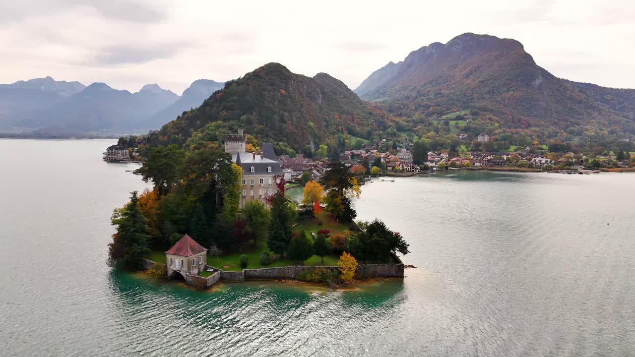 Château de Duingt, set on a walled headland jutting into the turquoise waters of Lake Annecy, while the village and steep wooded hills rise behind under an overcast Alpine sky, drone orbits