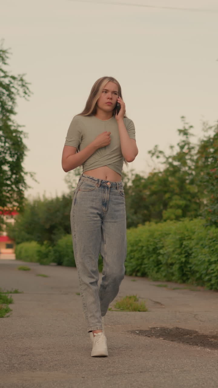 Young adult in casual jeans and a grey crop top walks on a paved path lined with lush green bushes and tall trees, engaged in a phone conversation, with one hand resting on her chest