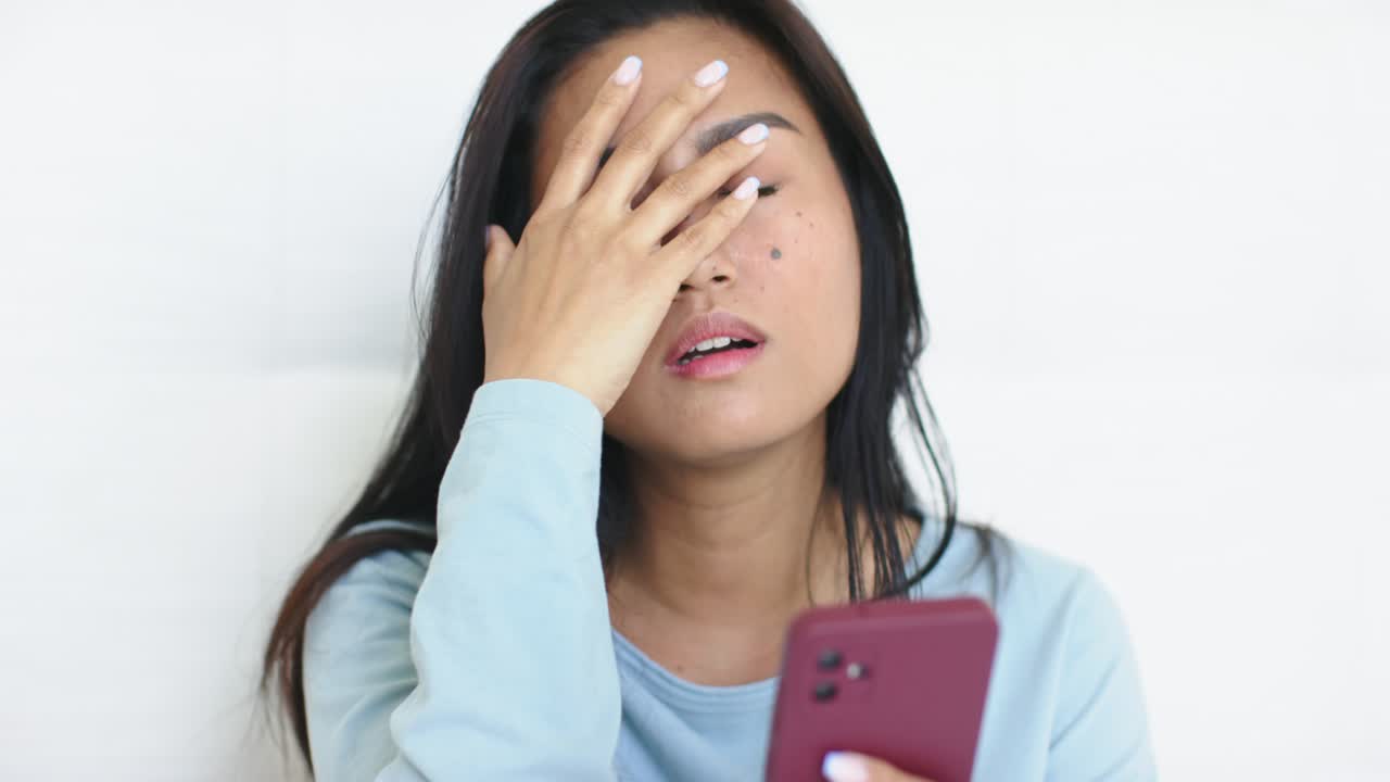 スマートフォンでメッセージを確認しながら額に手を当て、ソファに寄りかかるアジア人女性の画像テキスト