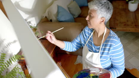 Focused-senior-biracial-woman-wearing-apron-and-painting-on-big-canvas-at-home,-slow-motion