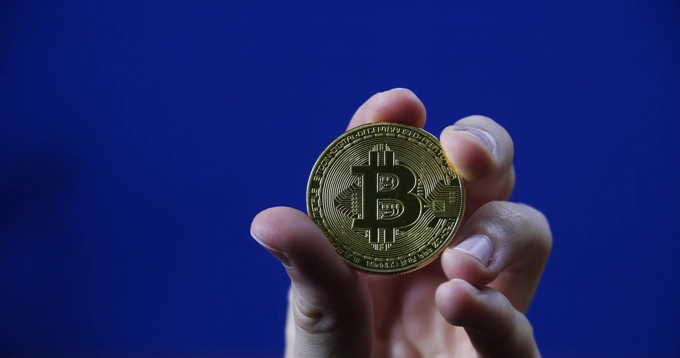 Uma mão segurando um Bitcoin dourado brilhante contra um fundo azul sólido em foco nítido.