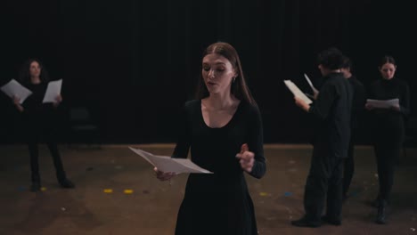 a confident young girl with glowing hair color in a black suit comes closer and reads out her text from a piece of paper while playing her role in the theater impression of emotions on stage during a rehearsal and preparation for a performance