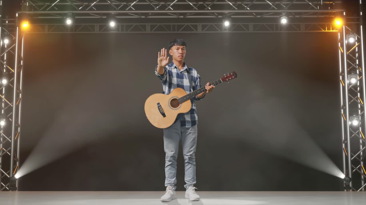 cuerpo lleno de adolescente asiático con guitarra haciendo gesto de parada mientras está de pie en el escenario