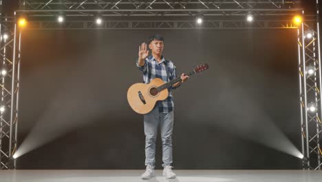 cuerpo lleno de adolescente asiático con guitarra haciendo gesto de parada mientras está de pie en el escenario