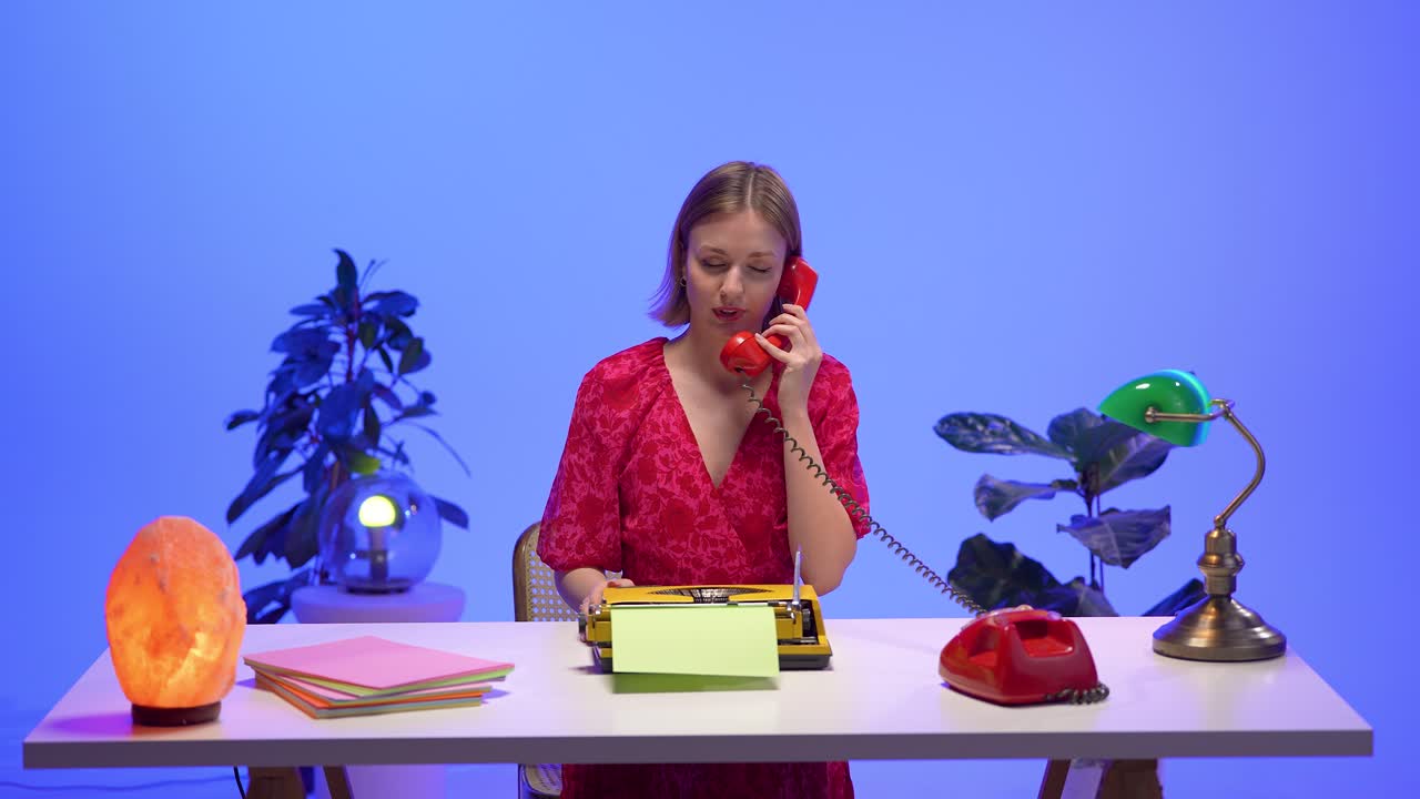 A woman in a red floral dress answers a classic red rotary phone while sitting at a bright desk with a yellow typewriter, pastel papers, and vintage lamps, surrounded by colorful retro decor
