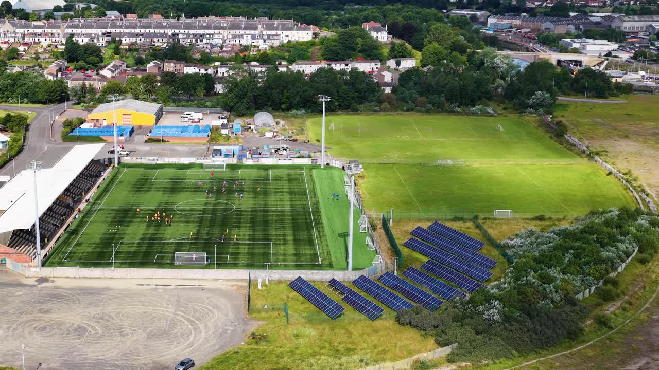 Drone flies toward football stadium with solar panels, training field, and green surroundings in daylight