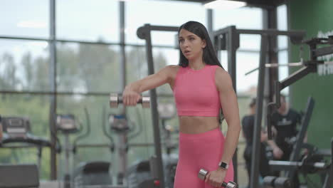 una mujer morena con un traje rosa eleva alternativamente las pesas a la parte superior delante de ella mientras entrena sus hombros en el gimnasio. ejercicio de pie para entrenar los hombros y los brazos. entrenamiento con pesas