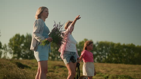 side view of family standing on hilly farmland under clear sky as mother points something interesting in distance to her daughter, while another woman stands nearby holding a bouquet of wildflowers