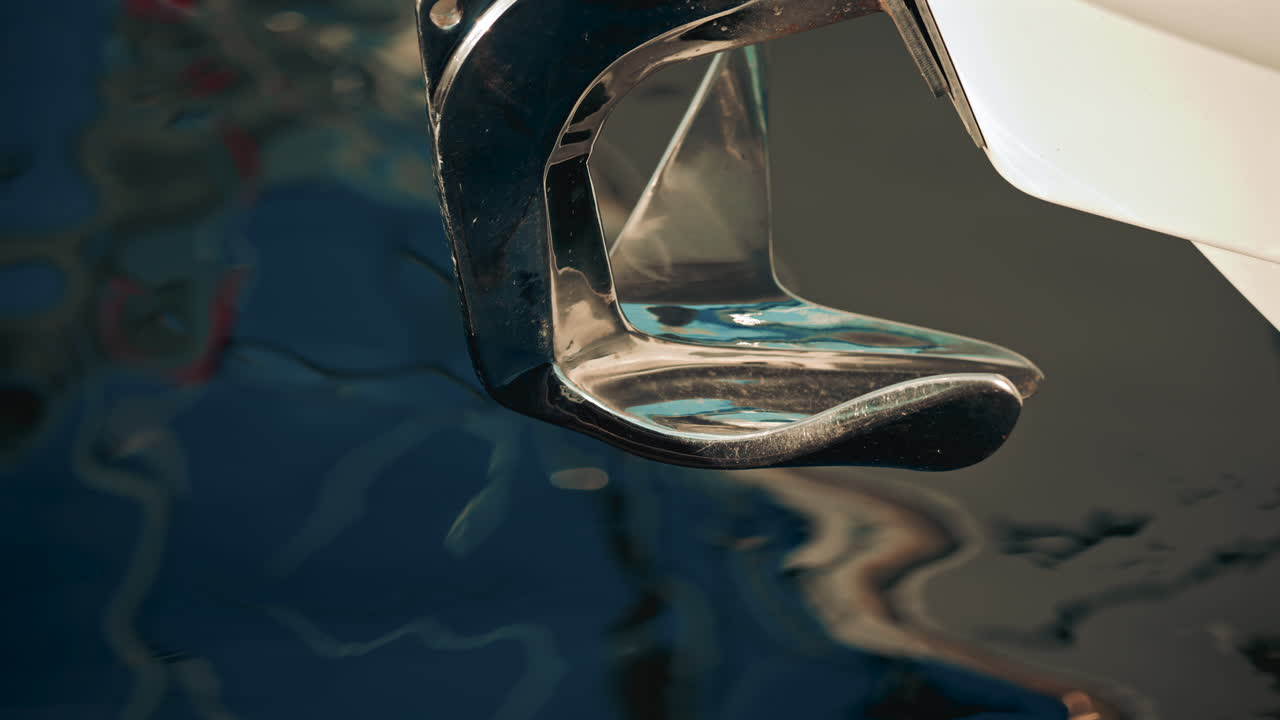 A chrome boat anchor with smooth curves, set against abstract blue and turquoise water reflections in a marina