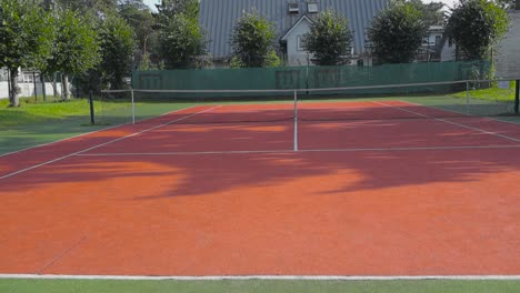 tennis court yard with a net in the middle during summer day time with sun shining and casting cinematic shadows. tennis field white lines are visible with trees in the background and green grass.