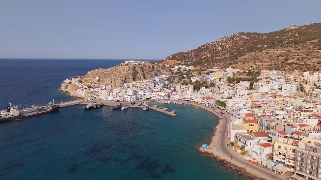 Stunning panoramic drone footage reveals the picturesque seafront and town of Pigadia, Karpathos, showcasing its beautiful coastline and vibrant harbor under a bright sunny sky