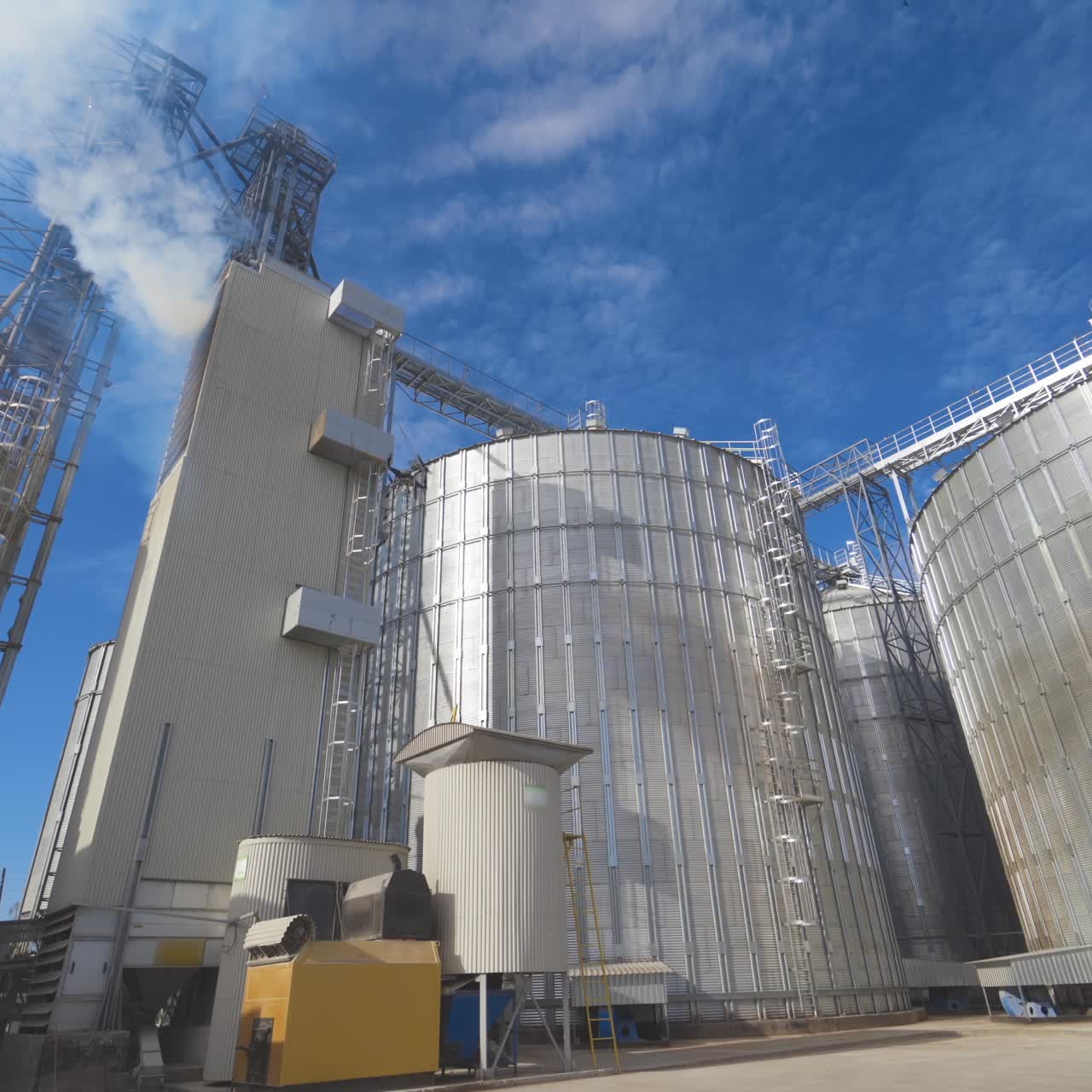 Agricultural Silos. Metal grain facility with silos. Storage and drying of grains, wheat, corn, soy, sunflower against the blue sky with white clouds