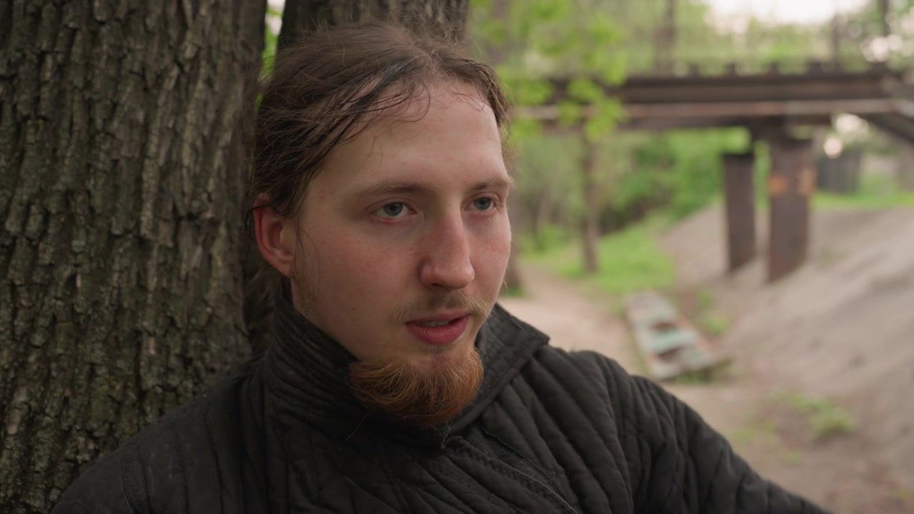 Primo piano di un uomo con barba rossa che indossa abiti imbottiti in stile medievale, che aggiusta e tiene il suo elmo nero mentre è in piedi vicino a un albero in un ambiente di parco all'aperto. La scena presenta una luce naturale soffusa.