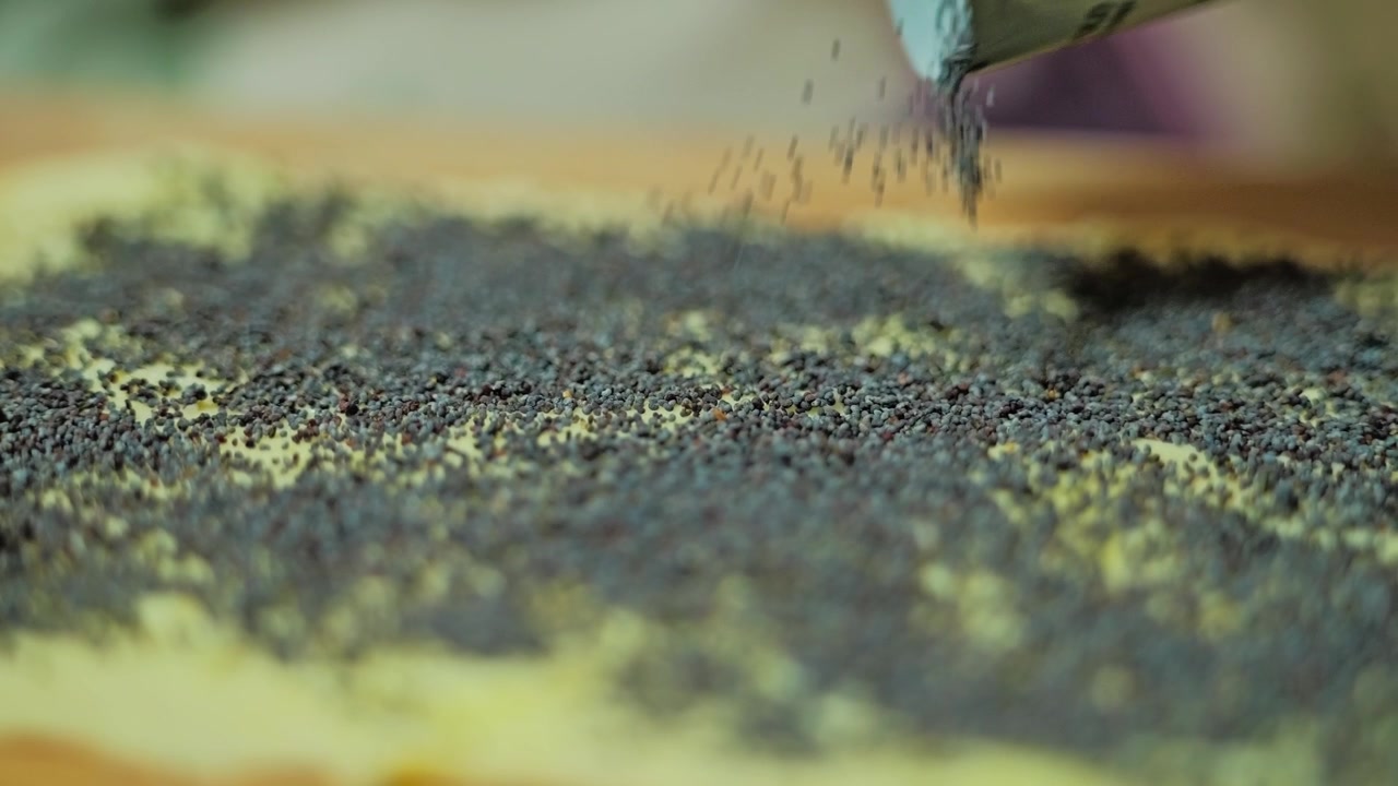 Close-up shows poppy seeds falling onto puff pastry dough in slow motion