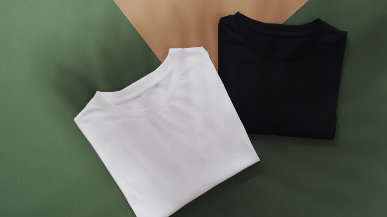 Video of flat lay of folded white and black t shirts with copy space on green and brown background