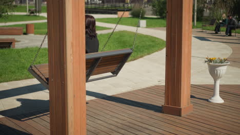 vista lateral de una mujer en una silla oscilante, su cabello soplando en el viento, en un tranquilo parque público con paisajismo detallado y personas en el fondo