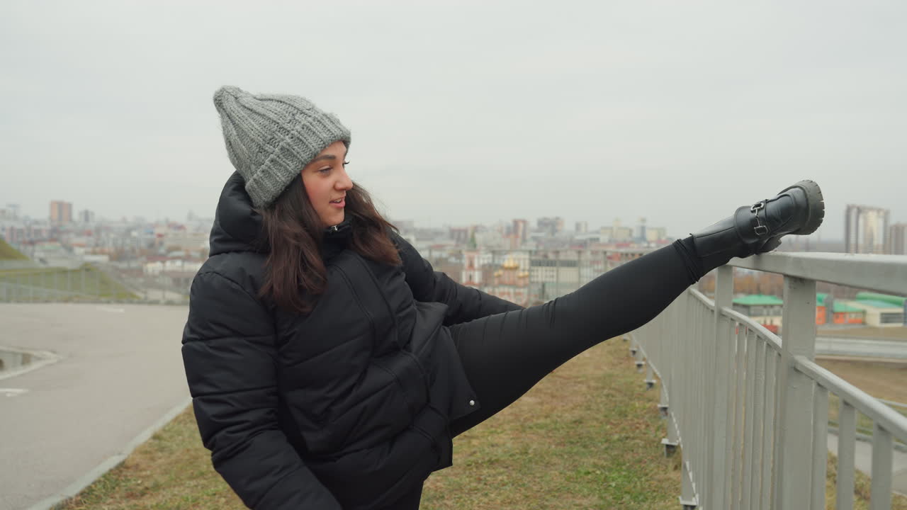 White woman performing high kick outdoors, winter coat and knit beanie, city skyline backdrop, upbeat dance warmup and playful rehearsal, arm rhythm and strong balance, urban overlook choreography