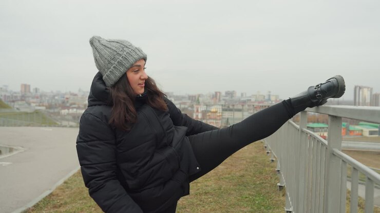 White woman performing high kick outdoors, winter coat and knit beanie, city skyline backdrop, upbeat dance warmup and playful rehearsal, arm rhythm and strong balance, urban overlook choreography