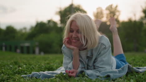 mom lying on picnic blanket in grass outdoors smiling heartfully, supporting head with hand, relaxed summer portrait, warm backlight creating soft glow, serene joyful moment