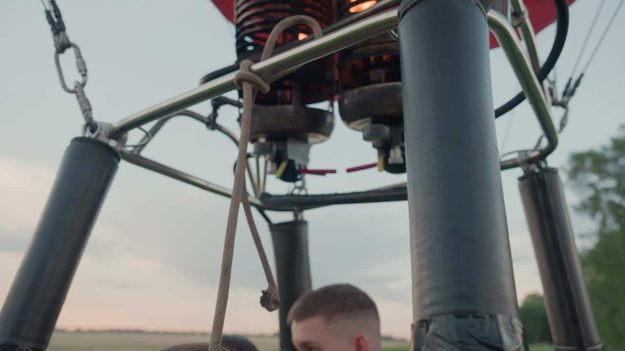people in wicker basket holding burner controls and tethers in farmland preparing launch of colorful hot air balloon at sunset amid open sky and grassy field before ascent adventure