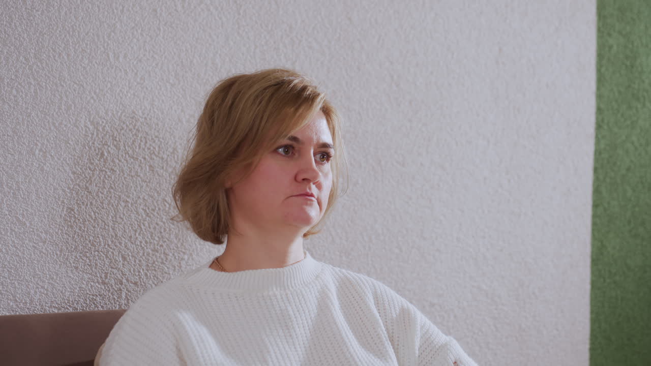 Reflective soul seated on sofa looking thoughtful against green accent wall wearing white sweater and neutral pants absorbing session atmosphere in modern therapy space during evening light