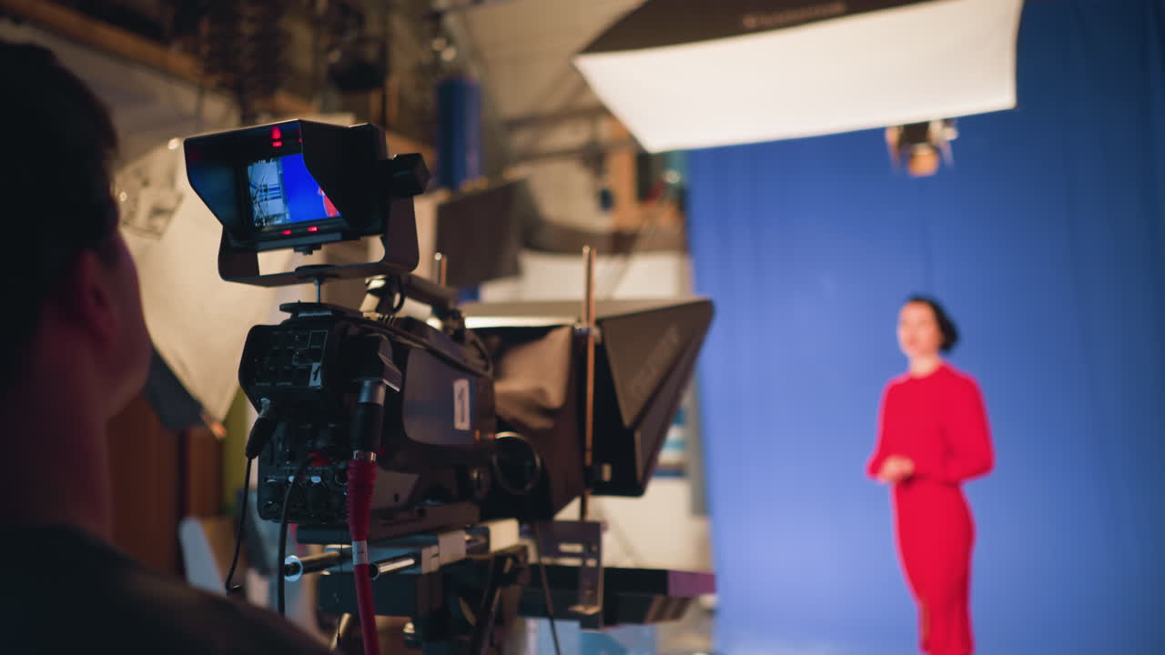 Cameraman filming female presenter in red dress standing in front of blue screen during professional studio shoot with teleprompter, camera monitor, lighting equipment, and production gear clearly visible in frame