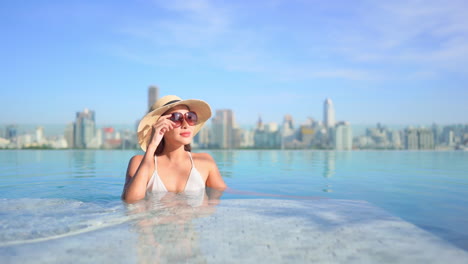 against the backdrop of an urban skyline, a pretty young woman lounges in a rooftop swimming pool