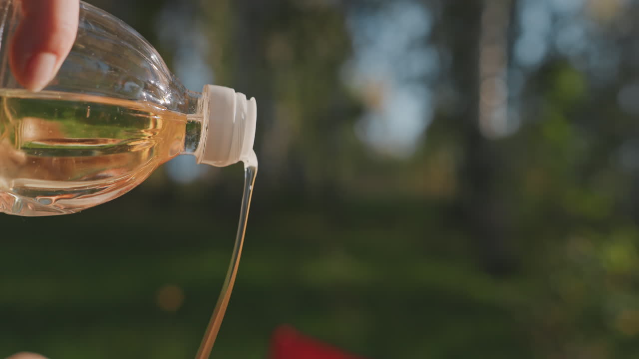Primo piano di olio versato lentamente in una padella da campeggio, flusso dorato scintillante da una bottiglia di plastica, mano che stabilizza il flusso, bokeh soffuso della foresta, scatto dettagliato in stile ASMR, camper bianco, focus sulla cucina sensoriale.