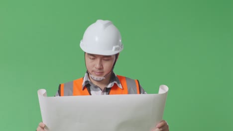 primer plano de ingeniero masculino asiático con casco de seguridad mirando el plano en sus manos y mirando a su alrededor mientras camina en el estudio de fondo de la pantalla verde