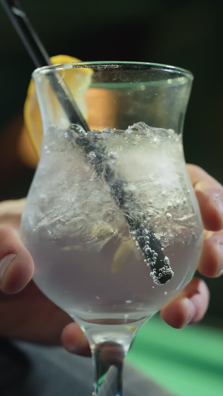Primo piano di un uomo, volto non visibile, che indossa una camicia bianca e pantaloni grigi e tiene in mano un bicchiere di limonata con una cannuccia nera. Cubetti di ghiaccio e una fetta di limone aggiungono dettagli rinfrescanti, creando un'atmosfera rilassata.