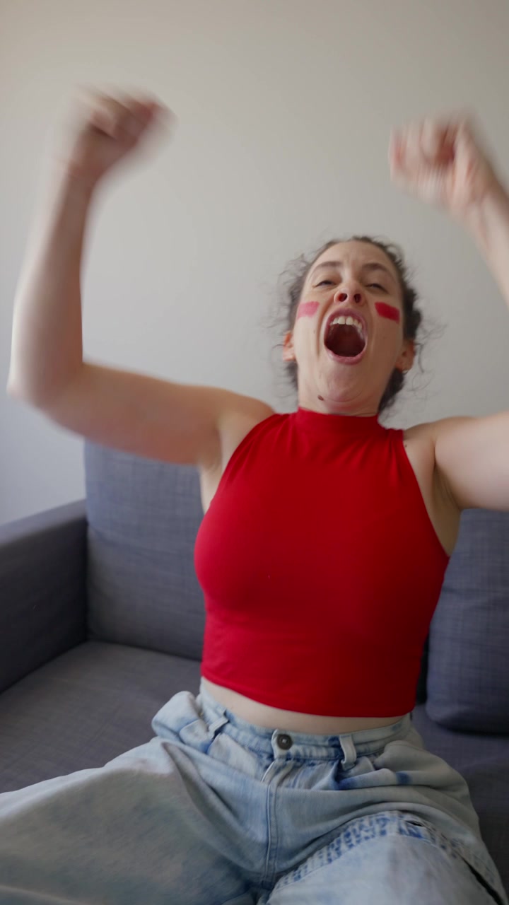 UGC vertical video - female sports fan screaming and celebrating win or score in living room watching live game on TV in her apartment or house. Passionate young woman red team supporter excited happy