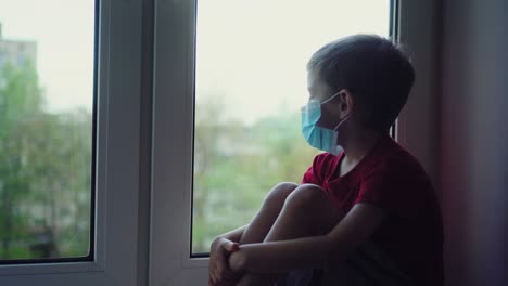 sad boy in medical mask sits on windowsill and looks out window, self quarantine 01