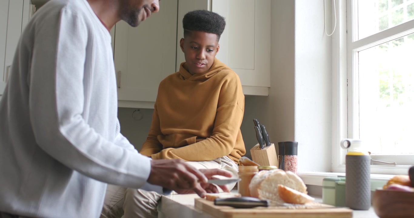 Uomo e ragazzo adolescente afroamericani che preparano uno spuntino, aprono un barattolo e spalmano la crema su un panino vicino alla finestra.