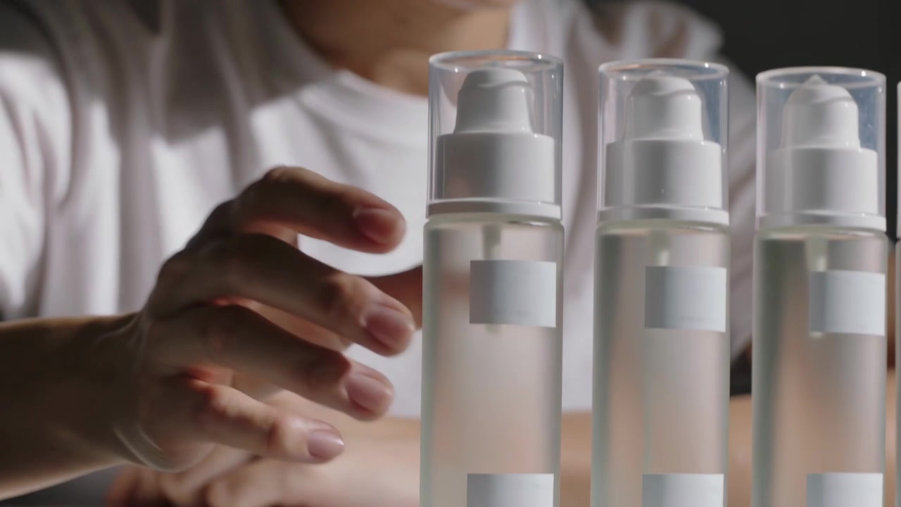 Luz suave iluminando mulher asiática de camiseta branca inspecionando e alinhando 3 frascos transparentes com bomba em uma mesa, organizando.