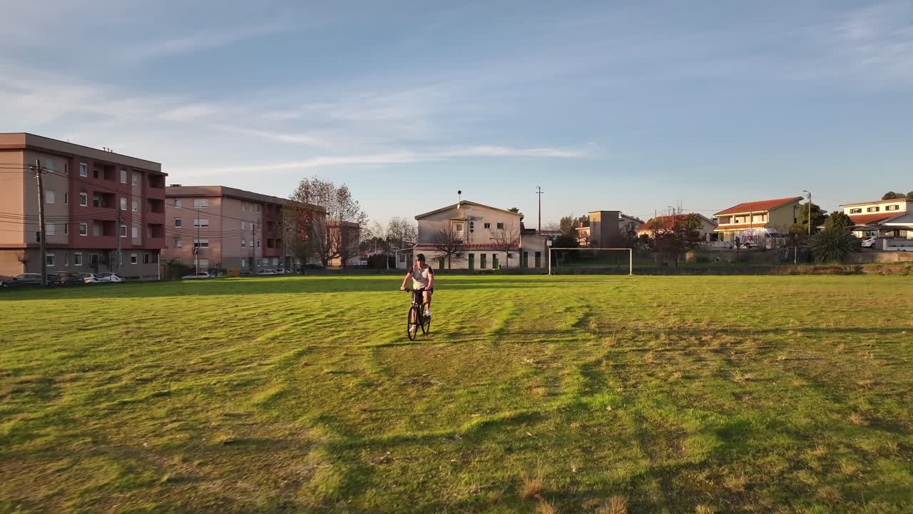 A drone flies backward as a cyclist rides towards it on a green field with a white goalpost. Small four-floor buildings are visible in the background, capturing a suburban scene.