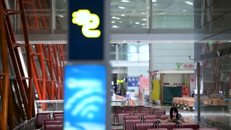 a female flight passenger sits and waits at the boarding gate in beijing international airport, using her smartphone, with a charging station sign in the foreground.