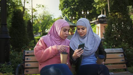 muslim ladies in colorful hijabs. they smiling, talking, enjoying coffee and looking at screen of mobile phone. sitting on bench in park. slow motion