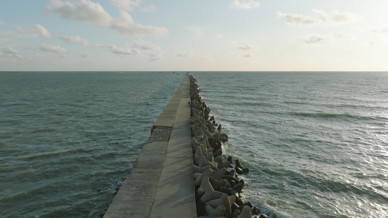 vista aérea del muelle de concreto del puerto de liepaja, costa del mar báltico, tarde soleada de verano, luz de la hora dorada, grandes olas salpicando, amplio disparo de drones avanzando bajo