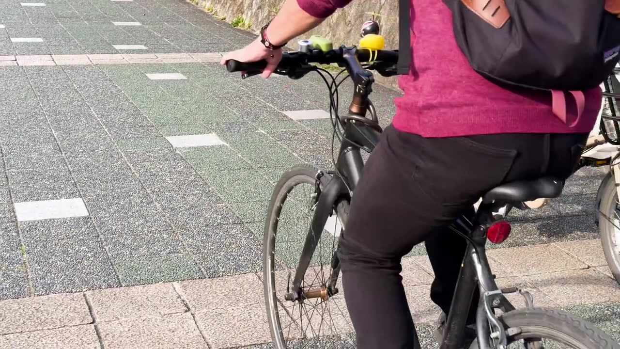 Commuters ride bicycles along a paved urban path beside a stone wall in bright daylight