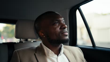 A-man-with-Black-skin-and-a-beard-in-a-brown-suit-rides-in-a-modern-car-and-mysteriously-looks-out-the-window-while-waiting-for-his-arrival-time.-Confident-male-businessman-driving-in-a-car-around-the-city